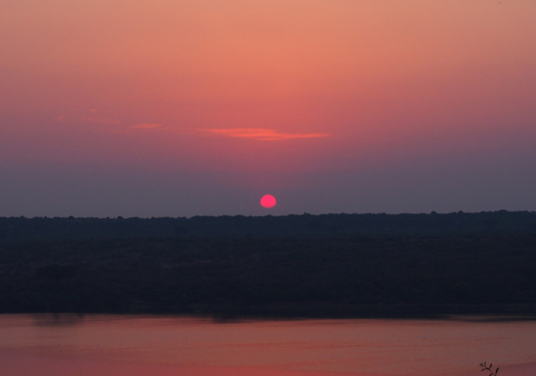 A bright pink sun hangs over a dark green jungle as it sets. The pink is reflected in the sky and the river below.