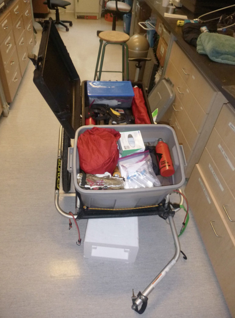 A bike trailer with two plastic totes filled with science lab equipment sits in a lab ready to go travelling.