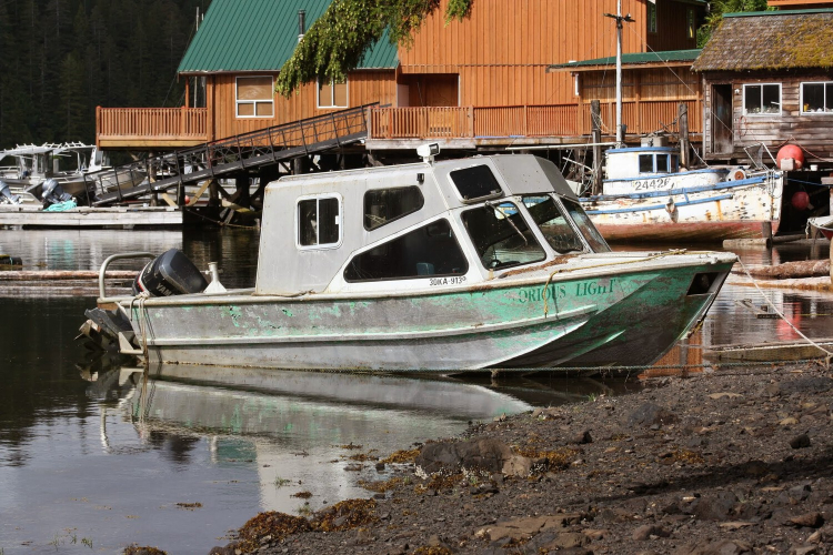A small weatherworn boat with an outboard motor is moored to shore with ropes. Several other fishing boats can be seen in the background.