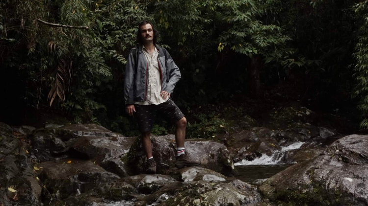 Pierre, wearing a beige shirt, gray jacket, black shorts, and hiking boots, stands on shiny dark brown rocks in a stream under a dense canopy of leaves.